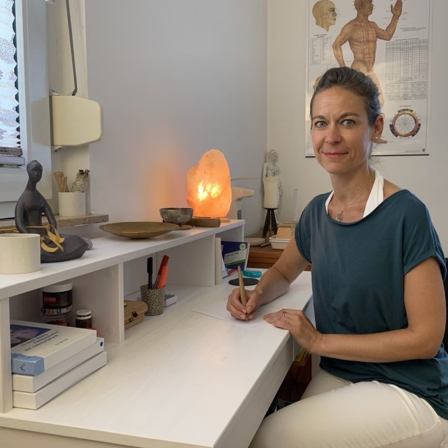 Heilpraktikerin Stephanie Bönisch in Praxis Heilpraktikerin Stephanie Bönisch sitzt an einem Schreibtisch mit orangefarbener Salzlampe und Meditationsfiguren.